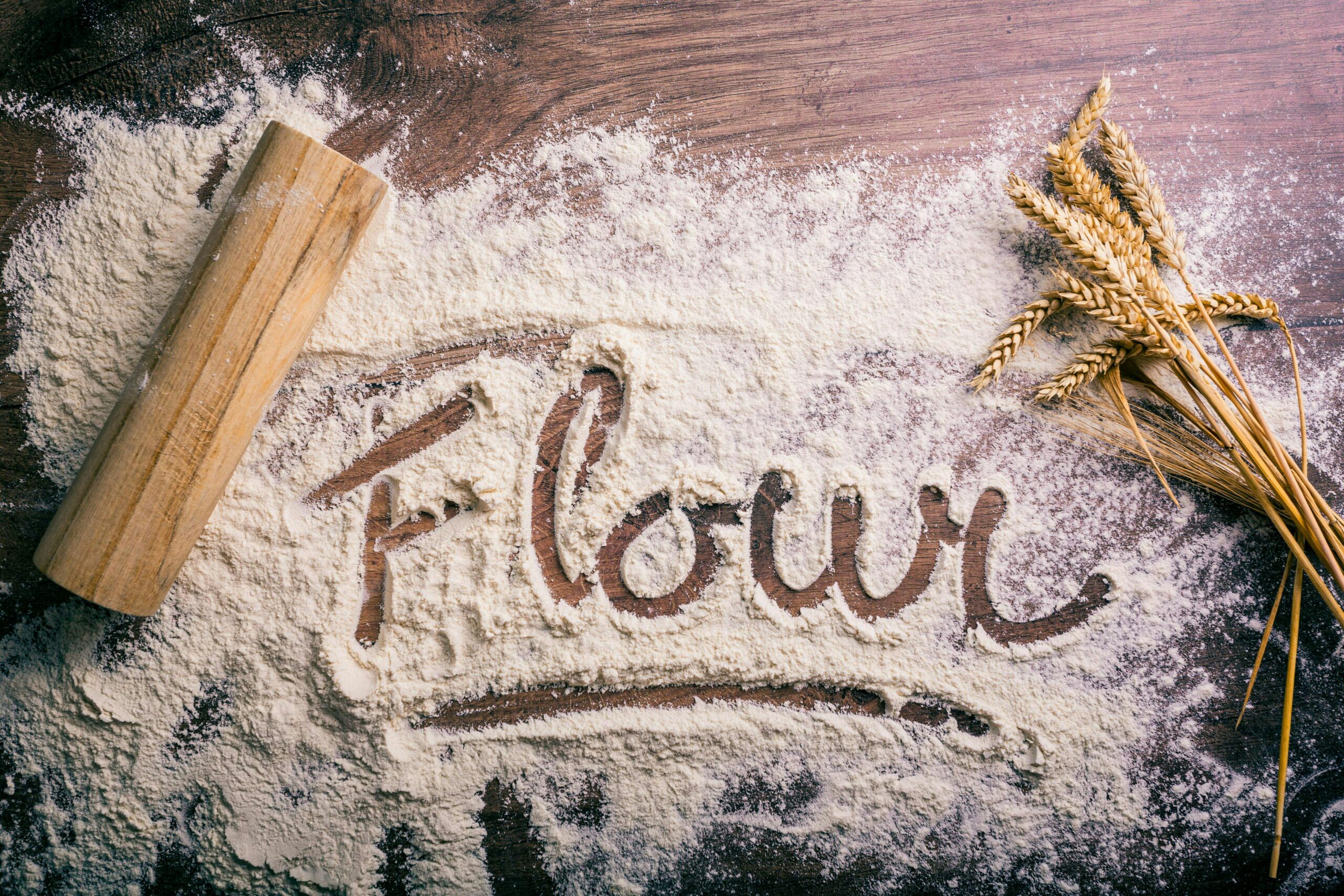 Flour scattered on a wooden table spelling 'flour,' with a rolling pin and wheat stalks nearby.